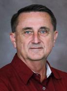 A man with short hair wearing a maroon shirt and white undershirt looks directly at the camera against a plain background.